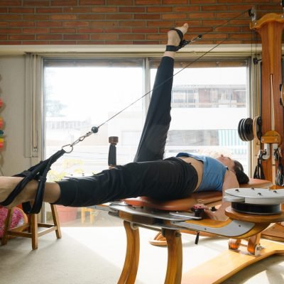 adult woman wearing blue sports clothing, lying on her back with one leg up and one down on pulley and weight machine, exercising muscles and working on joints at home